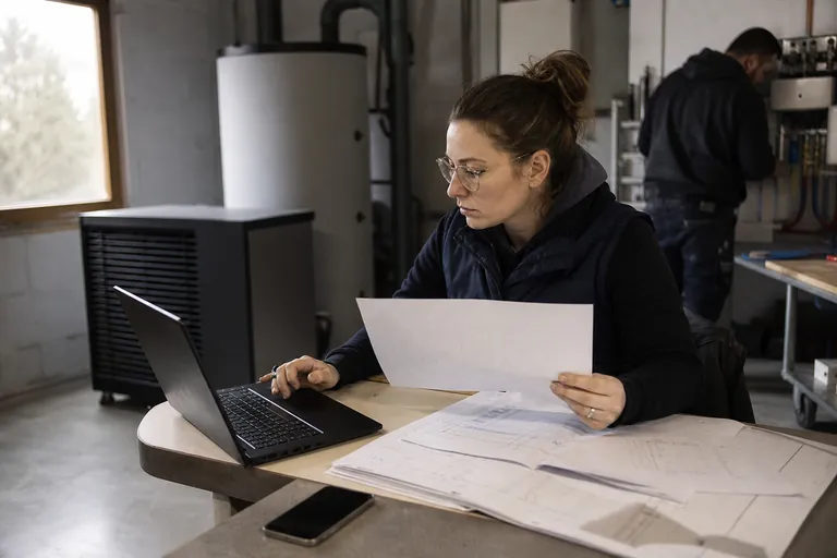 SHK Unternehmerin prüft eine Projektkalkulation auf einem Laptop neben technischen Unterlagen
